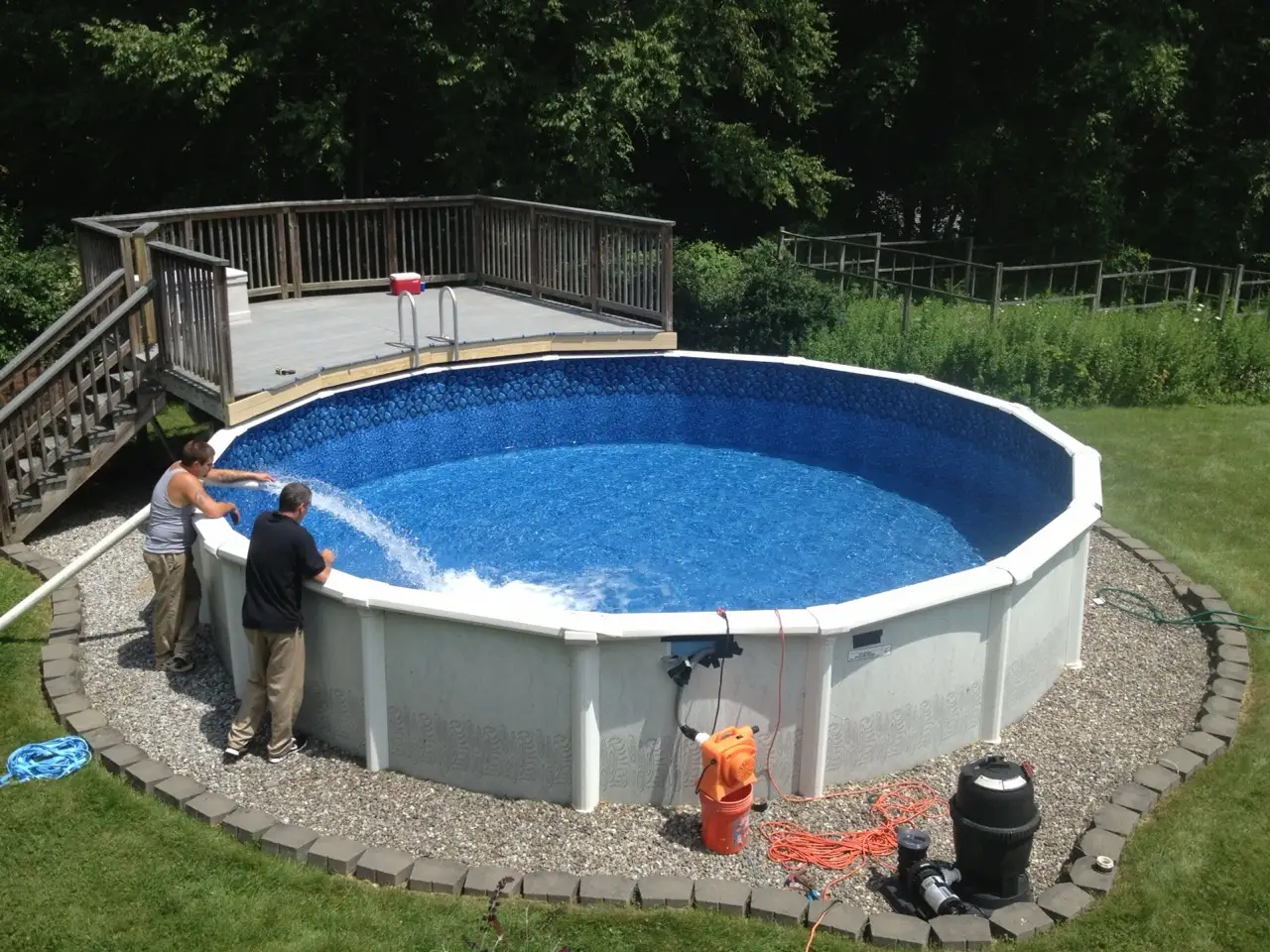 Two people filling a new round above-ground pool with a hose, beside a wooden pool deck