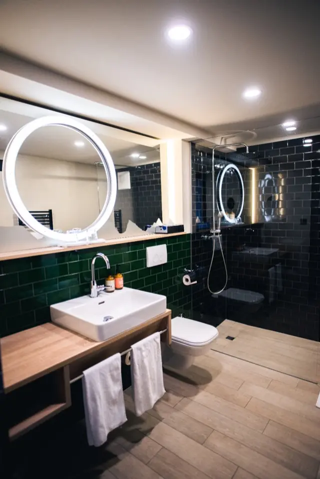 A remodeled bathroom with green and black tile, a floating wood vanity, a round lit mirror and a walk-in shower