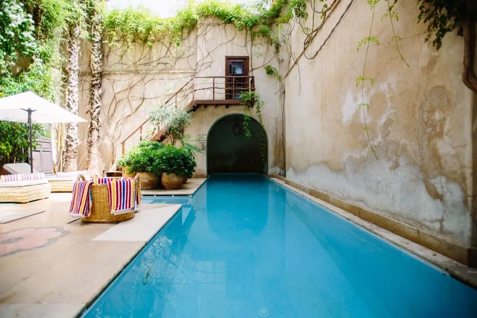 A long rectangular pool beside a stone patio with lounge chairs, an umbrella and potted plants