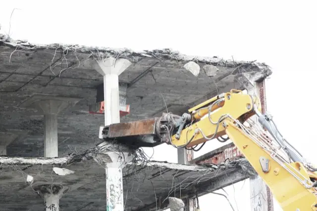 A yellow excavator arm tearing down a concrete floor slab during a demolition