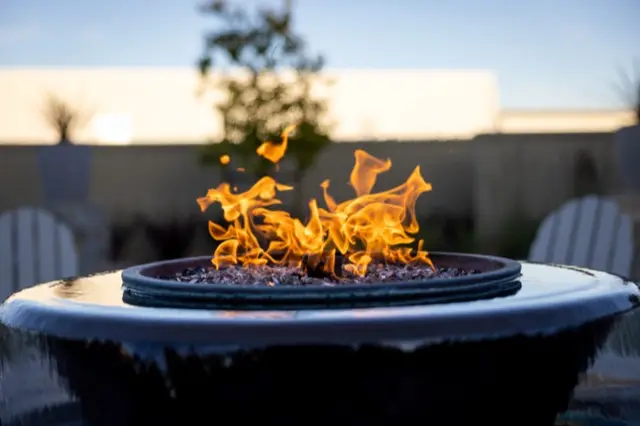 Flames rising from a round gas fire bowl on a patio at dusk, with outdoor chairs blurred behind