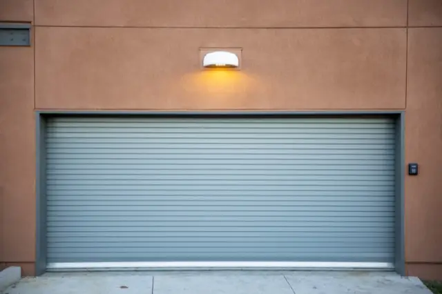 A closed grey roll-up garage door set in a tan stucco wall, under a single outdoor light