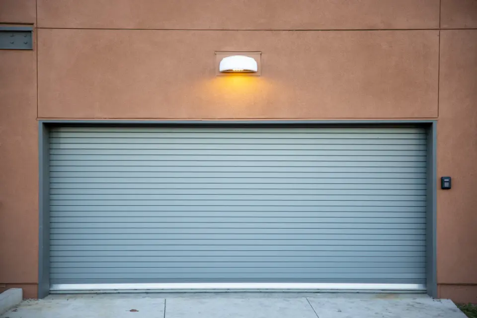 A closed grey roll-up garage door set in a tan stucco wall, under a single outdoor light