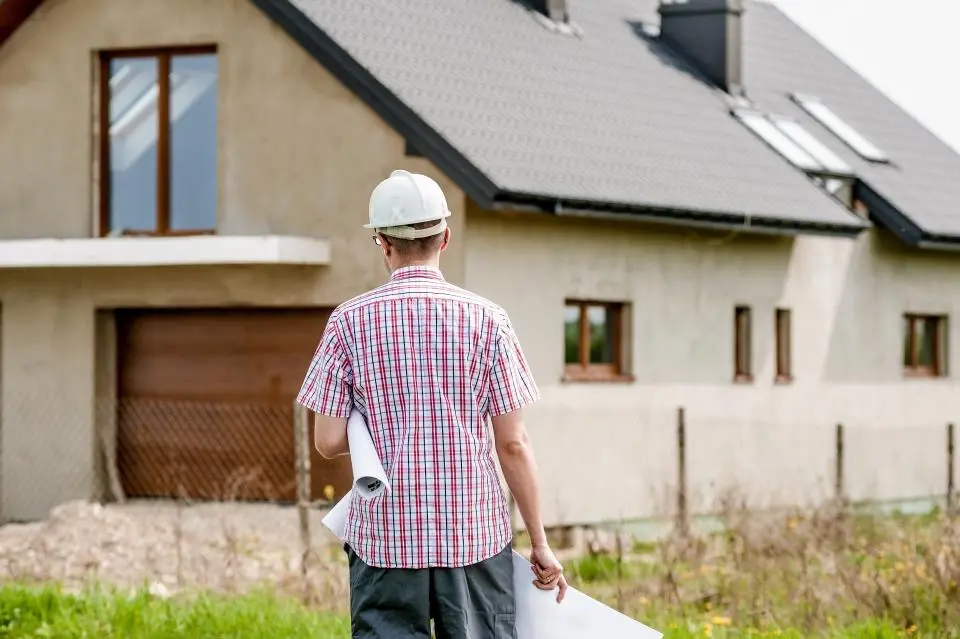 A man seen from behind carrying rolled-up plans toward a newly built house