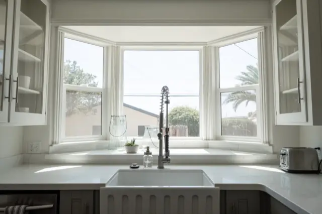 A white kitchen with a farmhouse sink, a tall faucet and a bay window above the counter