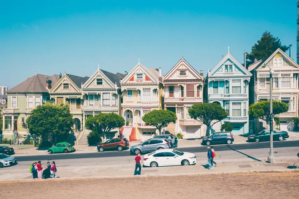 A row of painted Victorian houses facing a street with parked cars