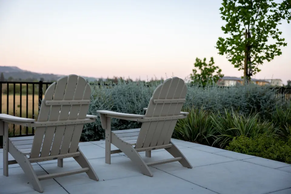 Two wooden Adirondack chairs on a concrete patio at dusk, with a garden and hills behind