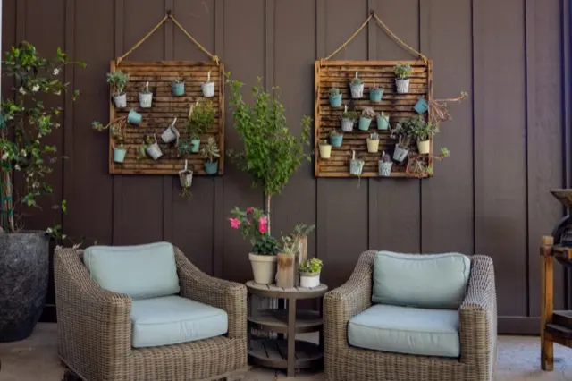 Two wicker armchairs with light blue cushions and potted plants on a patio against a dark board-and-batten wall