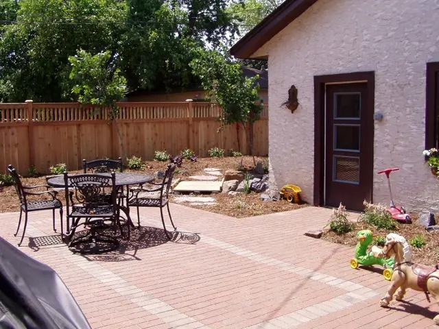 A brick paver patio with a metal table and chairs next to a white garden building and a wood fence