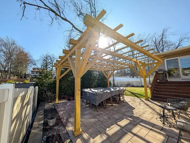 A freestanding wooden pergola over a paver patio with an outdoor dining table, sun shining through the rafters