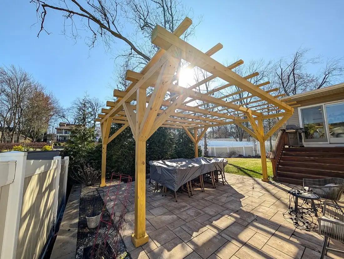 A freestanding wooden pergola over a paver patio with an outdoor dining table, sun shining through the rafters