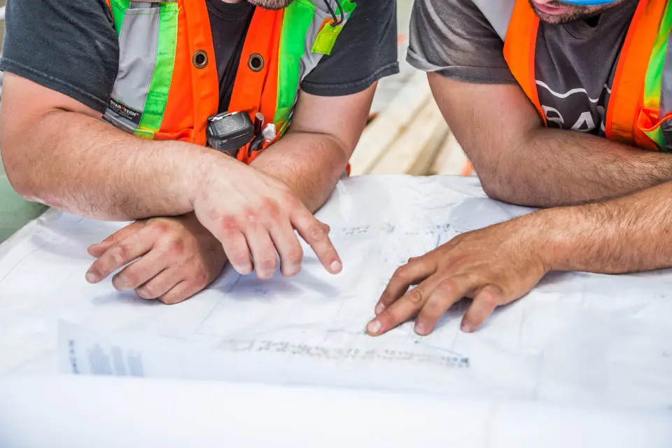 Two people in high-visibility vests pointing at construction drawings spread on a table