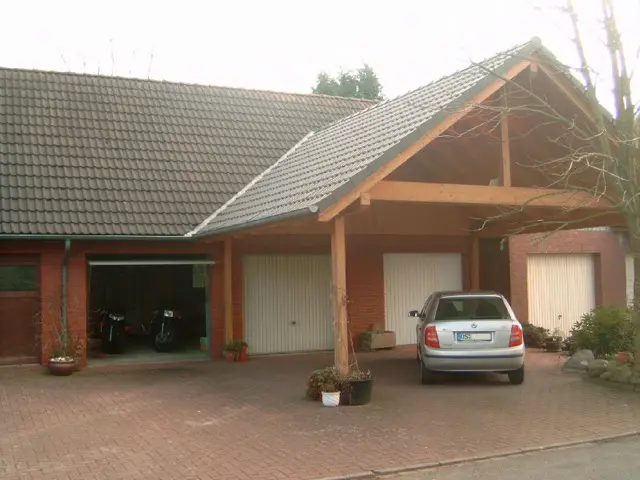 A gabled timber carport with a tiled roof sheltering a car in front of a row of garage doors